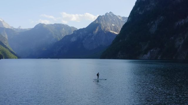 Aerial view Moving away shot, Man paddle boarding in the lake waves, Scenic view of the lake surrounded by mountains.