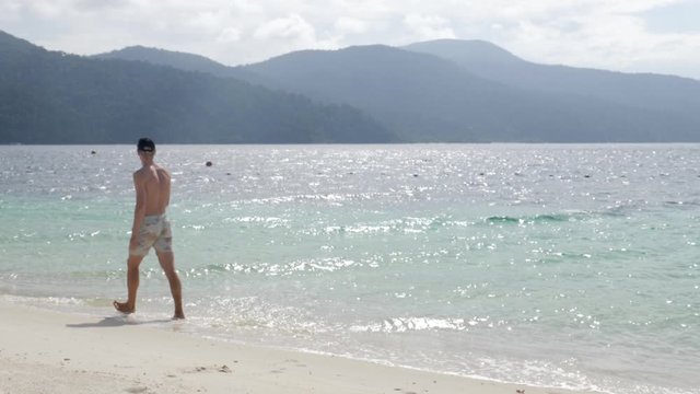 Ultra slow motion shot of young caucasian man walking relaxed with swimwear on stunning beach in Thailand