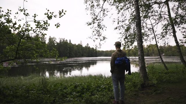 Young man with camera walks in forest and stops in front of lake, pan