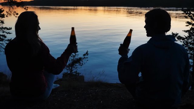 silhouettes of young couple clinking bottles of tasty beer sitting on bank of large calm river at sunset time backside view