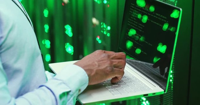 Close up of African American man among servers working on laptop computer, typing on keyboard and decrypting codes. Male analytic in data storage working on digital security and programming.