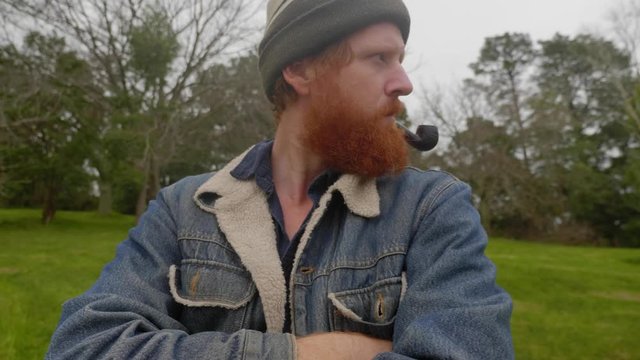 A shot of a bearded ginger mans face as he smokes from a tobacco pipe in the woods.