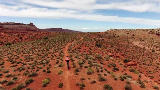 Aerial follow shot behind woman mountain biking on flat trail with red desert sandstone