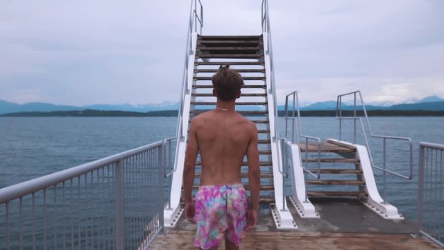 Slow motion shot of a young man walking towards diving board and walking up the stairs, watching over beautiful seascape.