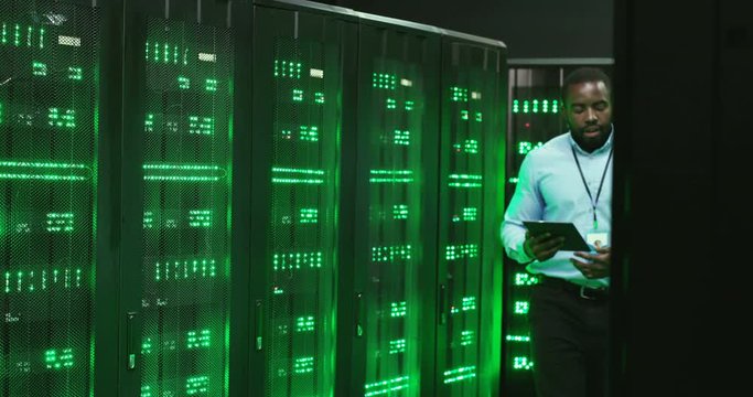 Mixed-races men walking together talking and watching data on tablet device and laptop computer. Software multiethnic engineers at servers room. Males analytics coworkers in datacenter discussing work