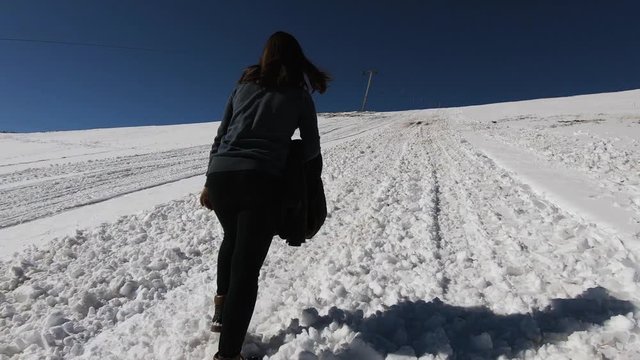 Girl slowly climbing up a snowy mountain. Shot from behind as she's walking along a ski slope, next to a ski lift. Blue skies and a snowy hill in the background.