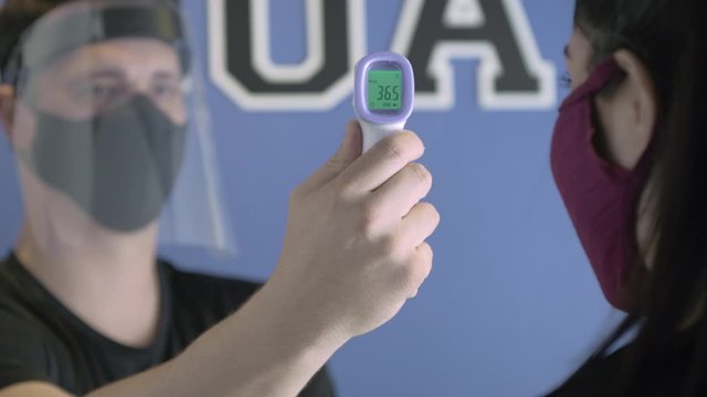 Health professional in mask and face shield  taking the temperature measure of a woman wearing a mask with a laser infra red thermometer.