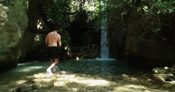 Young Caucasian shirtless and barefoot man wearing black bathing suit walks to fresh mountain pool by waterfall and dives in, low backward aerial