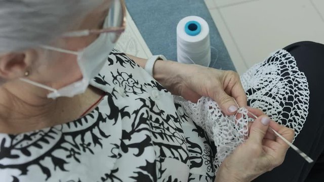 Old woman knitting with white thread and wearing mask on her face