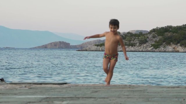 Caucasian toddler boy, running happily on a dick next to the sea, wearing a swimsuit, slow motion 120fps