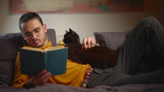 Cat lies on young man reclining in bed reading a book during pandemic