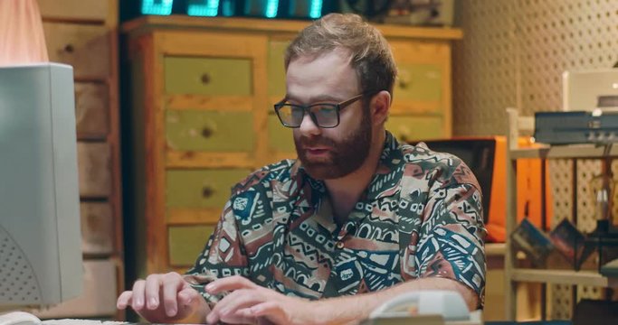 Caucasian young man in glasses and colorful shirt typing on keyboard while working at old computer. Portrait of old retro male programist or writer smiling to camera and giving thumb up. Vintage style