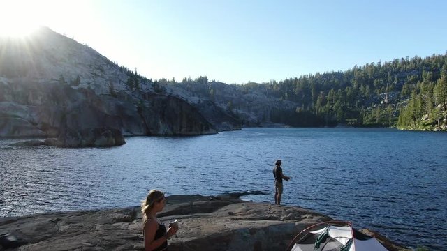 4K stabilized drone footage flying past a man swinging in a hammock during backpacking and hiking with family and friends in summer at an alpine lake fishing in California wilderness at sunset!