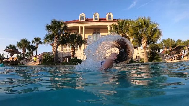 Young Caucasian girl standing whips and flings long hair in blue pool water splashing drops by tropical mansion home on blue sky sunny day, low vantage close up profile slow motion