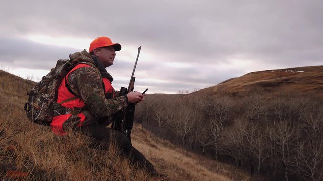 Hunter With A Rifle Sitting On A Grassy Hill Holding A Mobile Phone Searching Signal In A Hunting Field In Saskatchewan, Canada - Medium Shot