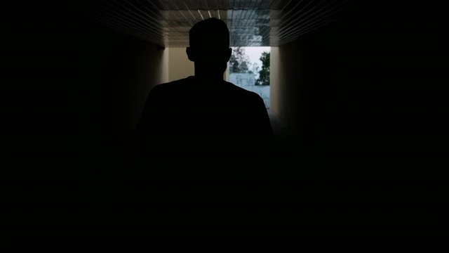 Silhouette of guy walks down dark corridor and sees light at the end of the tunnel. The man walks forward to the exit from the underpass on the highway.