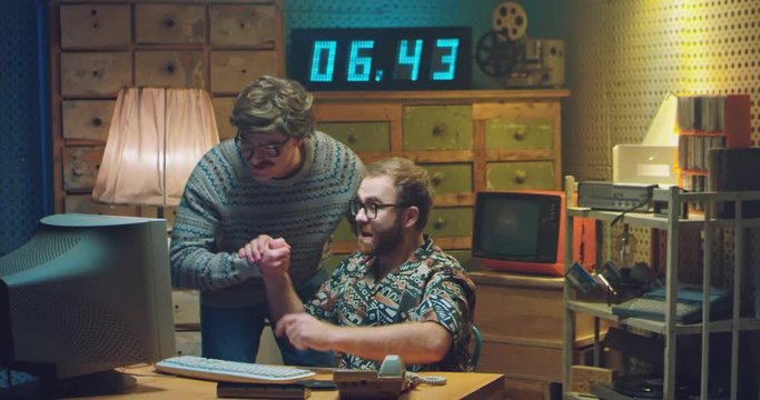 Two Caucasian males programists colleagues working at PC monitor and talking when deciding some problem. Retro style of 80's. Vintage computer. Men nerds of 70's. Help from friend. Giving five gesture
