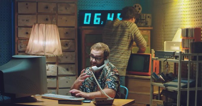 Two anxious Caucasian males programists in retro style room. Man in glasses answering vintage wired telephone ringing. Talking at computer monitor. Men nerds of 80's. Hackers of 70's in troubles.