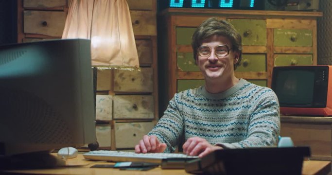 Portrait of Caucasian male nerd in glasses with mustache sitting at desk in retro room, smiling and working on computer. Man programist looking at camera. Vintage style of 70's. Gamer from 80's