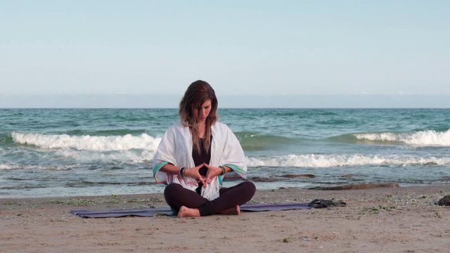 Young woman meditating while sitting on a yoga mat at the beach. Natural morning light wide shot with the Mediterranean Sea in the background.