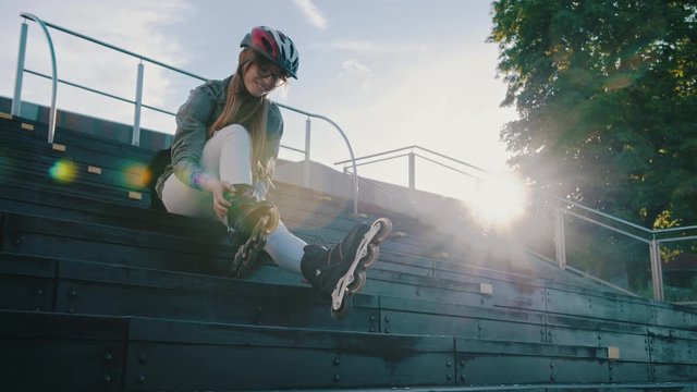 Young sporty woman adjusting rollerblades while sitting on the stairs in the skatepark. Direct sunshine, slow motion and low angle shot