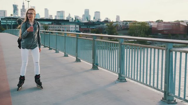 Excited young caucasian girl crossing the bridge on the rollerblades and using a smartphone