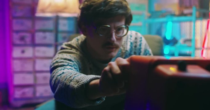 Caucasian funny young man nerd in glasses and with mustache sitting in retro style home and watching TV. Vintage television of 80's. Male standing and changing channels. Goofy guy of 90's.