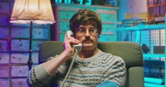 Caucasian young man nerd in glasses and with mustache sitting in retro style home and talking on wired vintage telephone of 80's. Angry male having phone call and putting. Goofy guy of 90's