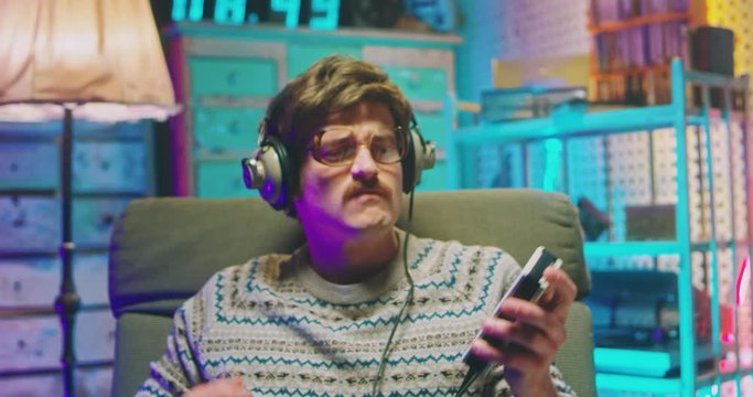 Young Caucasian man nerd in glasses, headphones and with mustache sitting in armchair in retro interior room and listening to music on vintage player. Male goofy imitating guitar play.