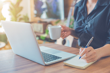 Woman is writing on a notebook with a pen and using a laptop to work in the office.