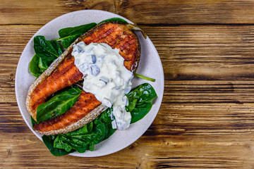 Plate with roasted salmon steak, tartar sauce and spinach leaves on a wooden table. Top view