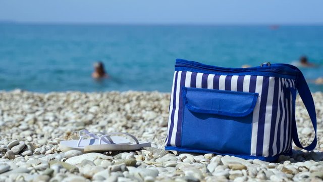 Beach bag and flip flops on pebbles with blue sea background and people swim on summer vacations