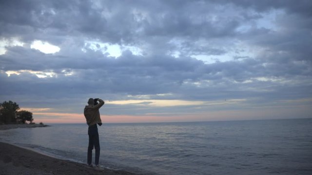 A Young Man Standing On The Shoreline While Taking Picture Of The Sunset Then Leaving During Early Evening - Wide Shot