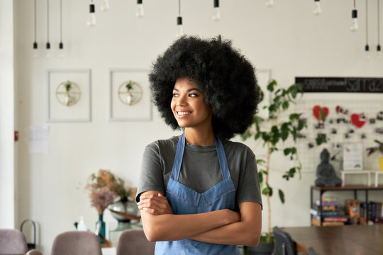 Smiling confident African American young woman with Afro hair modern cafe small business owner, female waitress in restaurant looking away standing arms crossed in cozy coffee shop interior. Portrait.