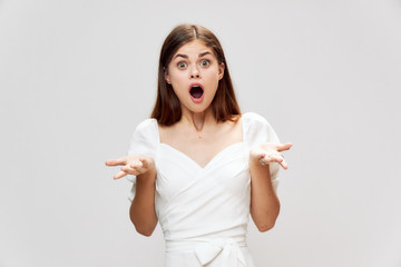 Surprised woman with wide open mouth spreads her hands in a white dress studio 