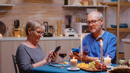 Old retired senior couple using phones in the kitchen. Sitting at the table browsing, searching, using smartphones, internet, celebrating their anniversary in the dining room.