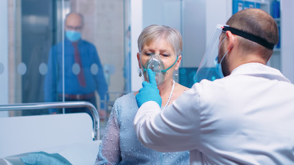 Doctor in mask and visor helping old lady to breathe with a respiratory oxygen mask while sitting...