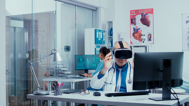 Senior Physician Using VR Glasses In Modern Private Clinic To Study Diseases In Virtual Space And Modern Technology. In The Background Modern Clinic With Glass Walls And Patients With Doctors In