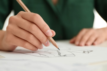 Woman drawing clothes with pencil on sheet of paper at white table, closeup