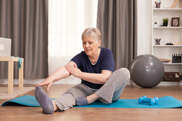 Active old woman exercising on the yoga mat in her comfortable apartment. Old person pensioner online internet exercise training at home sport activity with dumbbell, resistance band, swiss ball at