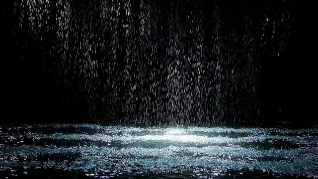 Spray spatters rain fall against on the water surface which glow. Black background. White neon lighting in dark studio. Slow motion.