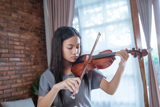 Asian girl playing the violin alone in the room against the curtain window backdrop