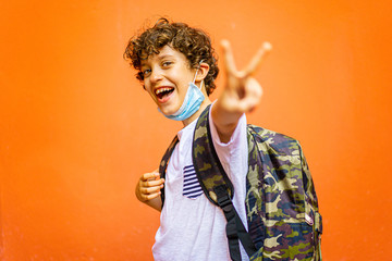 Happy schoolboy with a Covid-19, coronavirus, face mask making win or victory sign with his fingers. Isolated on a orange wall background.