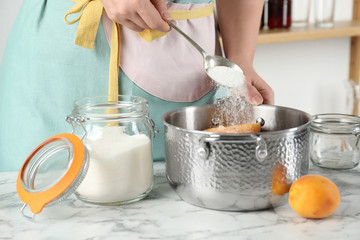 Woman adding sugar into pot with apricots at table in kitchen, closeup. Making delicious jam