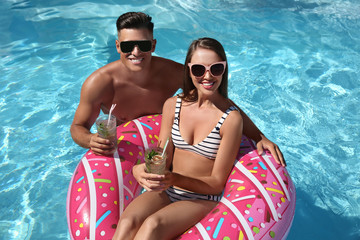 Happy couple with inflatable ring and cocktails in outdoor swimming pool on sunny summer day