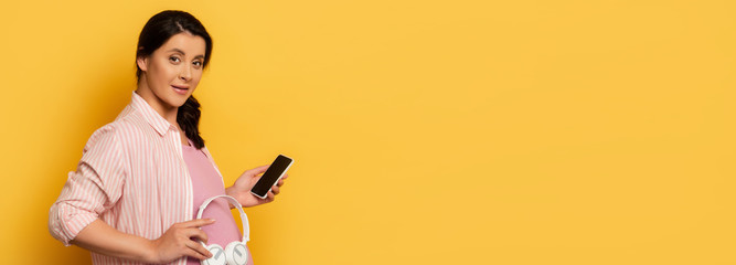 horizontal image of pregnant woman with smartphone holding wireless headphones near belly on yellow background