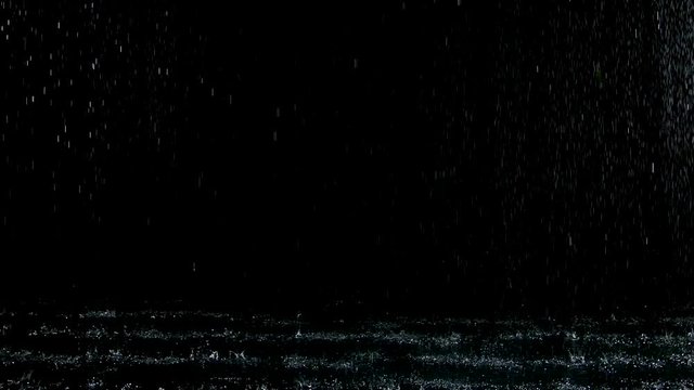From the ceiling spatters rain fall against on the water surface which glow. Black background. White neon lighting in dark studio. Slow motion. Close up.