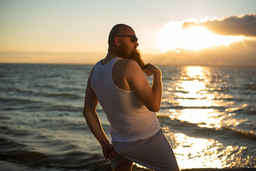 Fototapeta na wymiar A brutal bald man posing on the beach at sunset in his underwear and touching his red beard. A parody of a glamorous chick