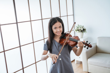 asian girl play the violin smiling while looking at the camera in the room against the window