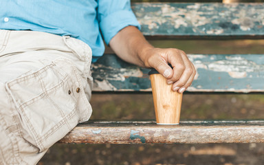 Cropped image of senior man sitting on park bench and holding reusable or easily recyclable bamboo cup for take away coffee. Tranquillity, relax and sustainable lifestyle concept.